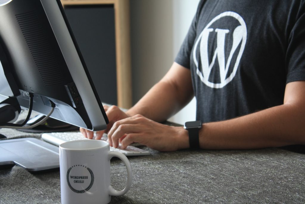 guy typing on desktop computer with wordpress logo on shirt and white mug on the table